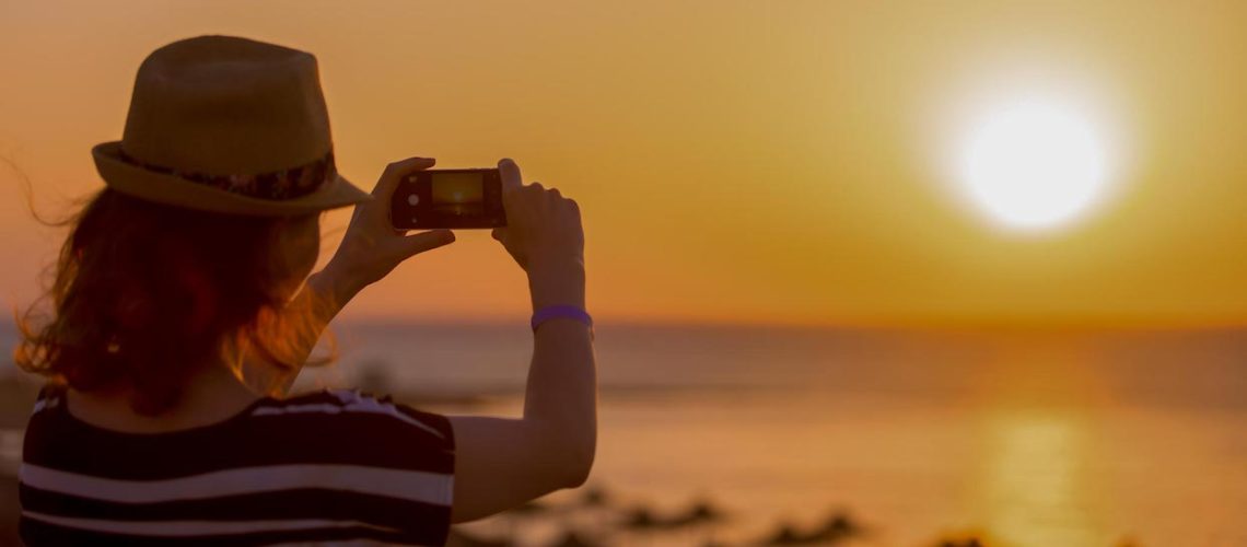 Young woman in straw hat and cute summer dress standing at the balcony with picturesque sea scenery, taking picture of sunset or sunrise on smartphone, back view